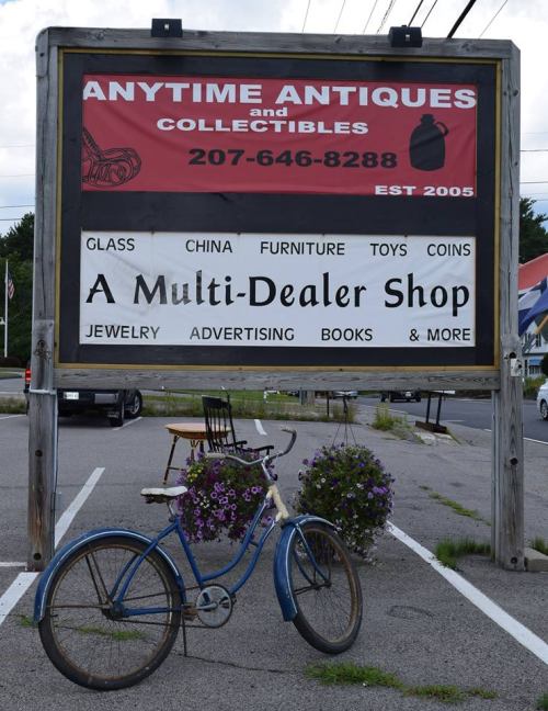 Sign for "Anytime Antiques and Collectibles" featuring a blue bicycle and flower pot, advertising various items for sale.