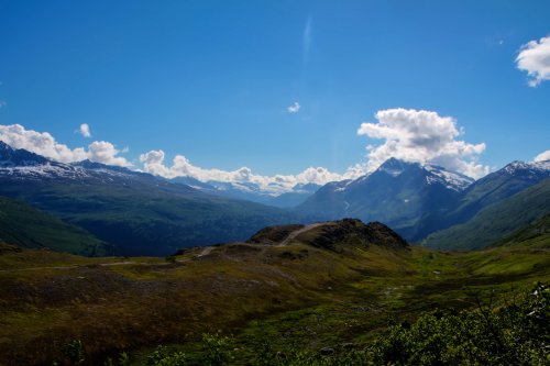 A scenic mountain landscape with green valleys, snow-capped peaks, and a clear blue sky dotted with clouds.