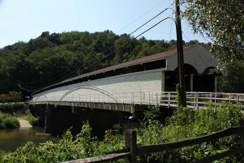 A covered bridge spans a river, surrounded by lush greenery and trees, with power lines overhead.