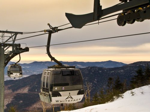 Two gondolas on a ski lift against a backdrop of mountains and a sunset sky. Snow covers the ground below.