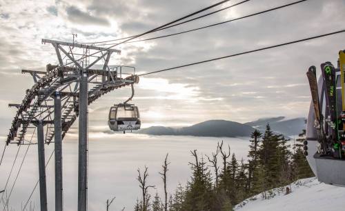 A ski gondola ascends a mountain, surrounded by clouds and trees, with skis leaning against a nearby structure.