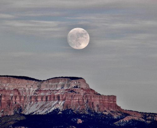 A full moon rises above a rugged mountain landscape, partially obscured by clouds.
