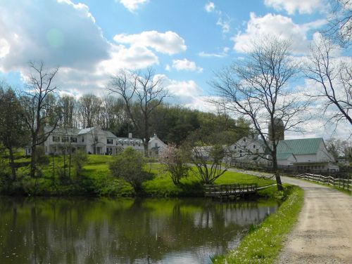 A serene landscape featuring a pond, a winding path, and a white house surrounded by trees and blue skies.