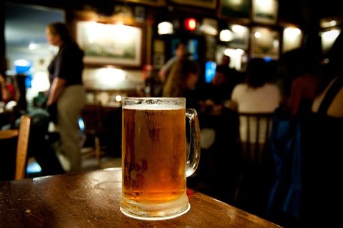 A frosty mug of beer sits on a wooden table in a dimly lit bar, with patrons mingling in the background.