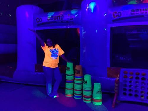 A person in a bright yellow shirt poses next to colorful stacking cups in a dimly lit, neon-themed play area.