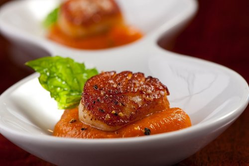 Seared scallop on a bed of vibrant orange sauce, garnished with a fresh green leaf, served in a white dish.