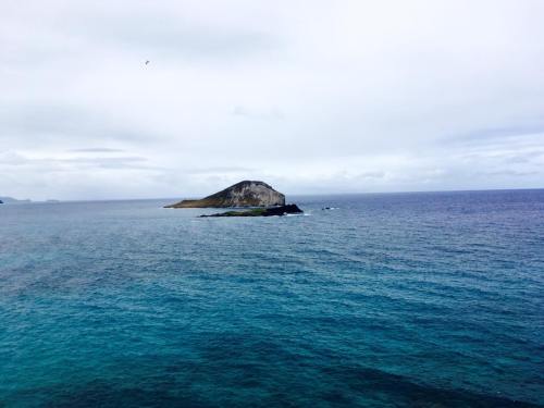 A small island rises from the calm blue ocean under a cloudy sky. Waves gently lap at its rocky shore.