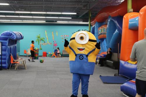 A person in a Minion costume stands in a colorful indoor play area with inflatable structures and children playing.