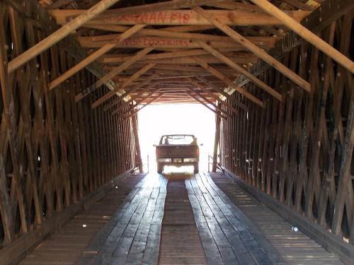 A vehicle drives through a wooden covered bridge, illuminated by light at the far end.