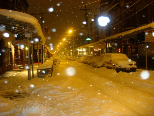 A snowy street at night, with cars covered in snow and streetlights glowing through falling snowflakes.