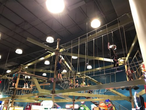Children navigate a rope course in an indoor amusement center, surrounded by bright lights and arcade games below.