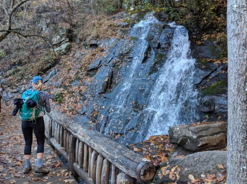 A hiker stands by a wooden railing, admiring a small waterfall cascading over rocky terrain surrounded by autumn leaves.