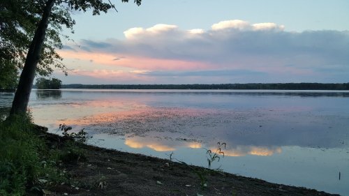 A serene lakeside view at sunset, with colorful clouds reflecting on the water's surface and trees lining the shore.
