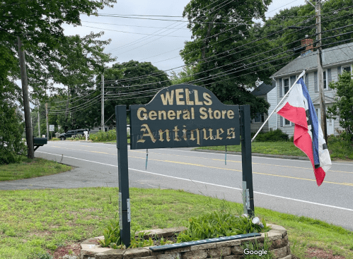 Sign for Wells General Store and Antiques, with a flag nearby, set along a quiet road surrounded by greenery.