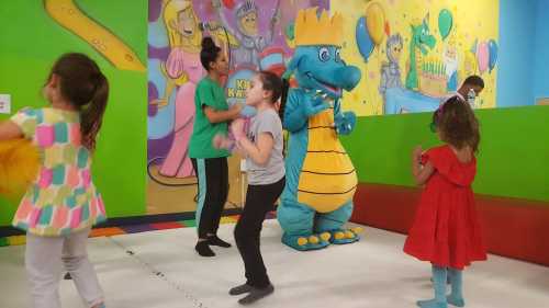 Children dancing with a blue dinosaur mascot in a colorful play area, with balloons and party decorations in the background.