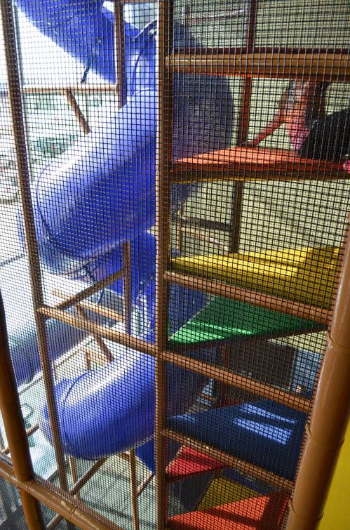 Colorful indoor playground featuring a blue slide and multi-colored stairs, viewed through a safety net.