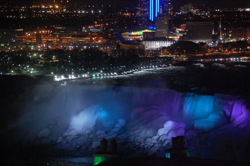 A nighttime view of illuminated Niagara Falls with colorful lights and a city skyline in the background.