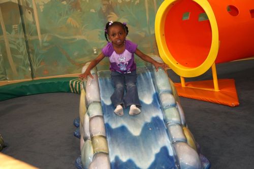 A young girl in a purple shirt sits on a colorful slide in a playful indoor environment.