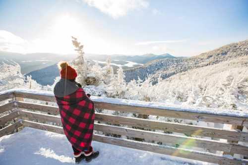 Person in a red and black checkered blanket stands on a snowy balcony, overlooking a winter landscape with mountains.