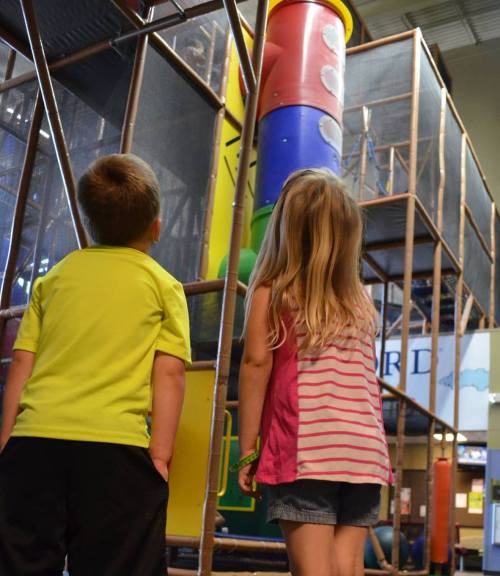 Two children stand in front of a colorful indoor play structure, looking up in awe.