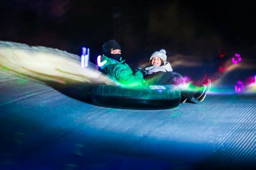 Two people joyfully tubing down a snowy slope, surrounded by colorful lights at night.