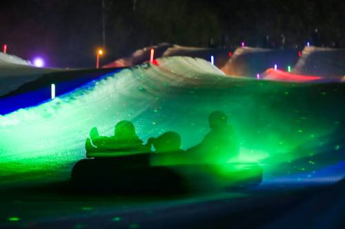 Two people on a snow tube glide down a brightly lit slope with colorful lights at night.
