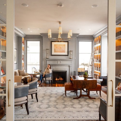 A cozy library with a fireplace, bookshelves, and a woman sitting in an armchair, surrounded by elegant decor.