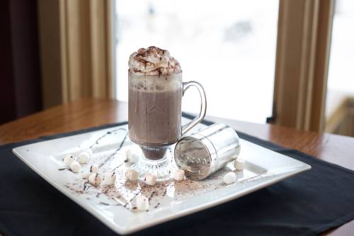 A glass of hot chocolate topped with whipped cream and cocoa, surrounded by mini marshmallows on a decorative plate.