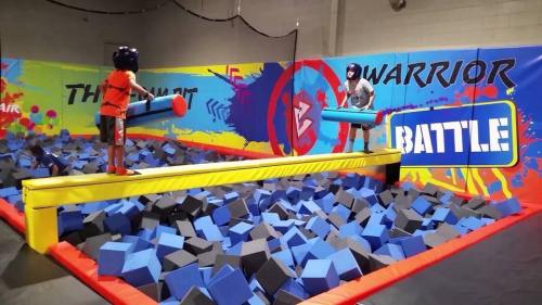 Two kids in helmets stand on a balance beam over a foam pit, preparing to battle with padded poles in a colorful arena.