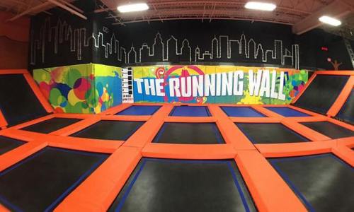 A vibrant indoor trampoline park featuring a colorful wall with "The Running Wall" text and multiple trampolines.
