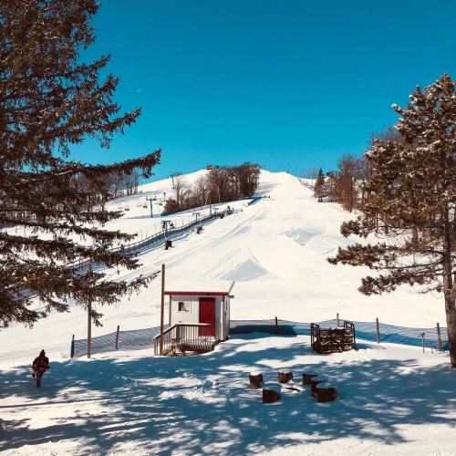 A snowy ski slope with a clear blue sky, surrounded by trees and a small building in the foreground.