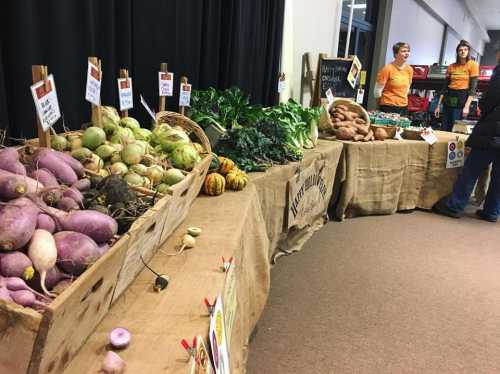 A market stall displays various fresh vegetables, with two vendors in orange shirts engaging with customers.