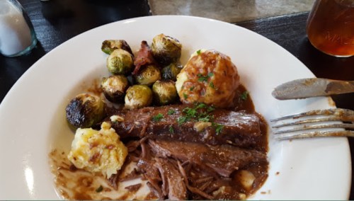 A plate of tender beef with gravy, roasted Brussels sprouts, and mashed potatoes, garnished with herbs.
