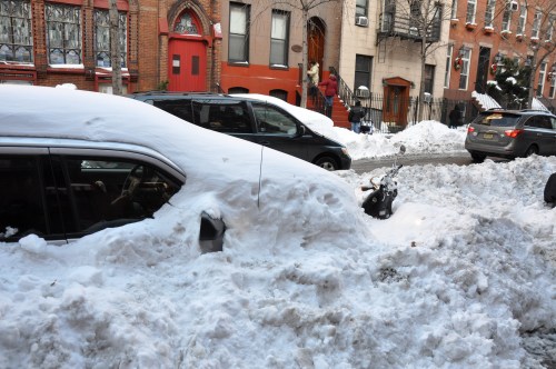 A car almost completely buried in snow on a city street, with other vehicles and buildings in the background.