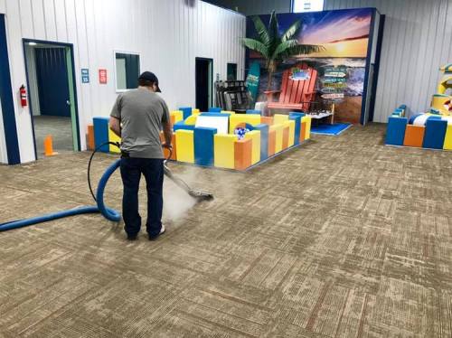 A person cleans a carpet with a steam cleaner in a colorful indoor space with beach-themed decor.