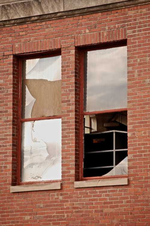 Two broken windows in a brick wall, reflecting a cloudy sky and partially obscured interior.