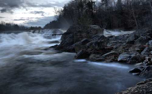 A turbulent river flows over rocks under a cloudy sky, surrounded by trees and mist.