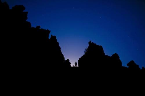 Silhouette of a person standing between dark rock formations against a twilight sky filled with stars.