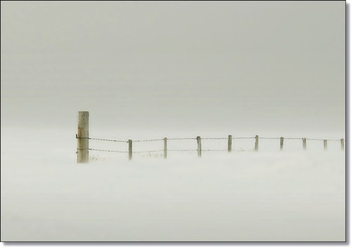 A lone fence post stands in a foggy landscape, partially obscured by a thick layer of mist.
