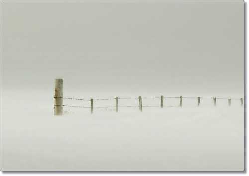 A lone fence post stands in a foggy landscape, partially obscured by a thick layer of mist.