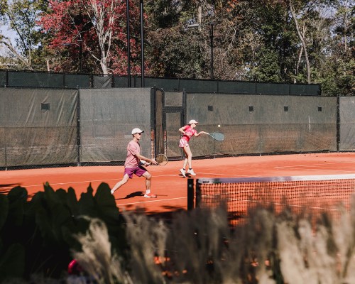 Two tennis players in pink outfits rally on a clay court surrounded by greenery and trees.