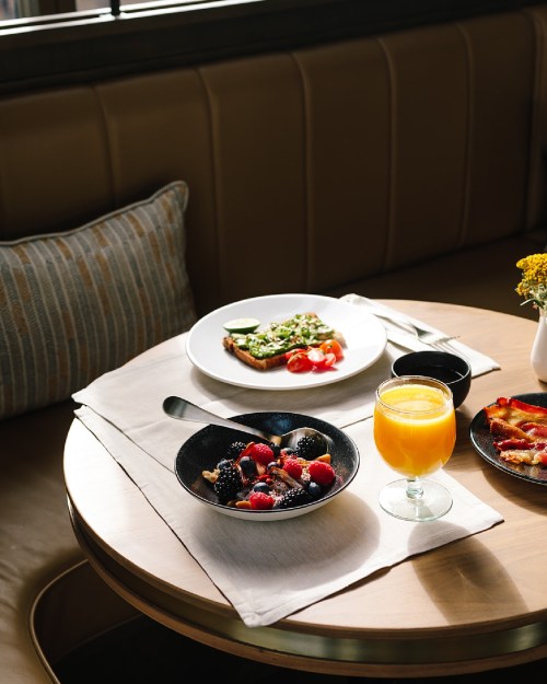 A cozy table setting with a fruit bowl, toast with greens, and drinks, surrounded by soft seating and flowers.