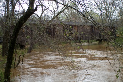 A wooden covered bridge spans a river, surrounded by trees and muddy water, under a cloudy sky.