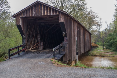 A wooden covered bridge spans a river, surrounded by greenery, with a gravel road leading to its entrance.