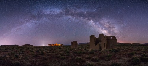 A panoramic view of a starry night sky with the Milky Way arching over ruins in a grassy landscape.