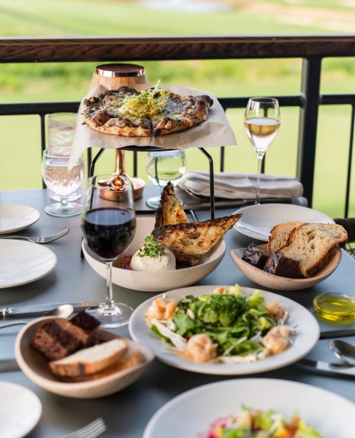 A beautifully arranged table with gourmet dishes, including pizza, salad, bread, and wine, set against a scenic backdrop.