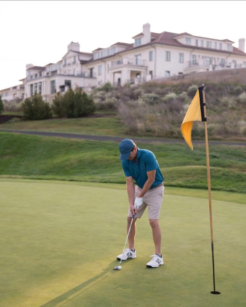 A golfer in a blue shirt and shorts putts on a green, with a yellow flag and a large house in the background.