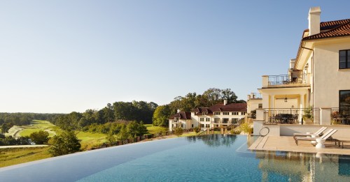 A luxurious infinity pool overlooks a scenic landscape and elegant villas under a clear blue sky.