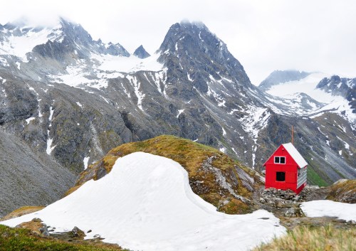 A small red cabin sits on a rocky hillside, surrounded by snow-capped mountains and a cloudy sky.