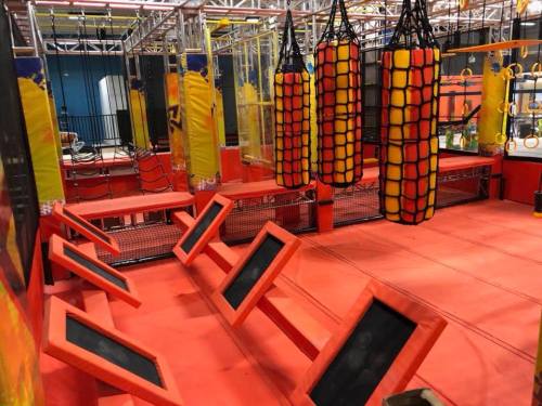 An indoor obstacle course featuring red and yellow climbing structures, hanging bags, and padded flooring.